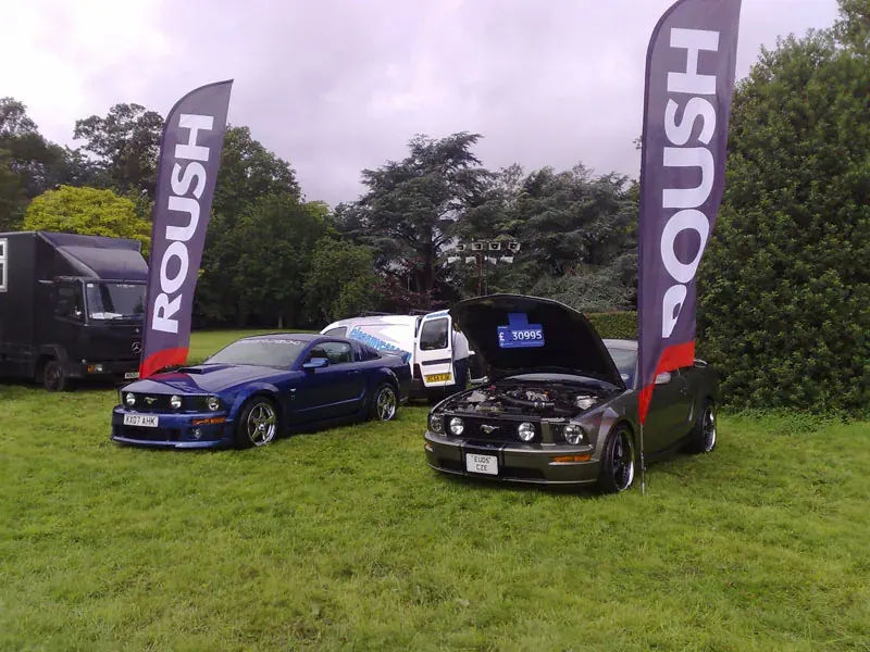 Two Mustangs displayed beneath Roush flags at an outdoor UK car show